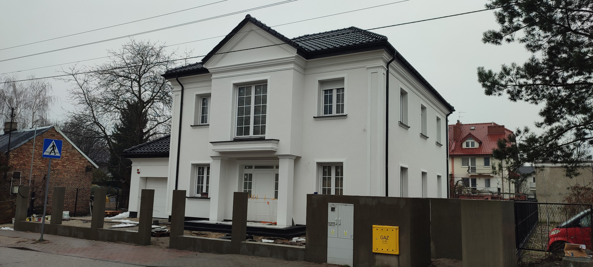 Elewacja dwukondygnacyjnego, białego domu z czarnym dachem, widoczna skrzynka gazowa i fragment ogrodzenia. W tle inne budynki i drzewa bez liści.