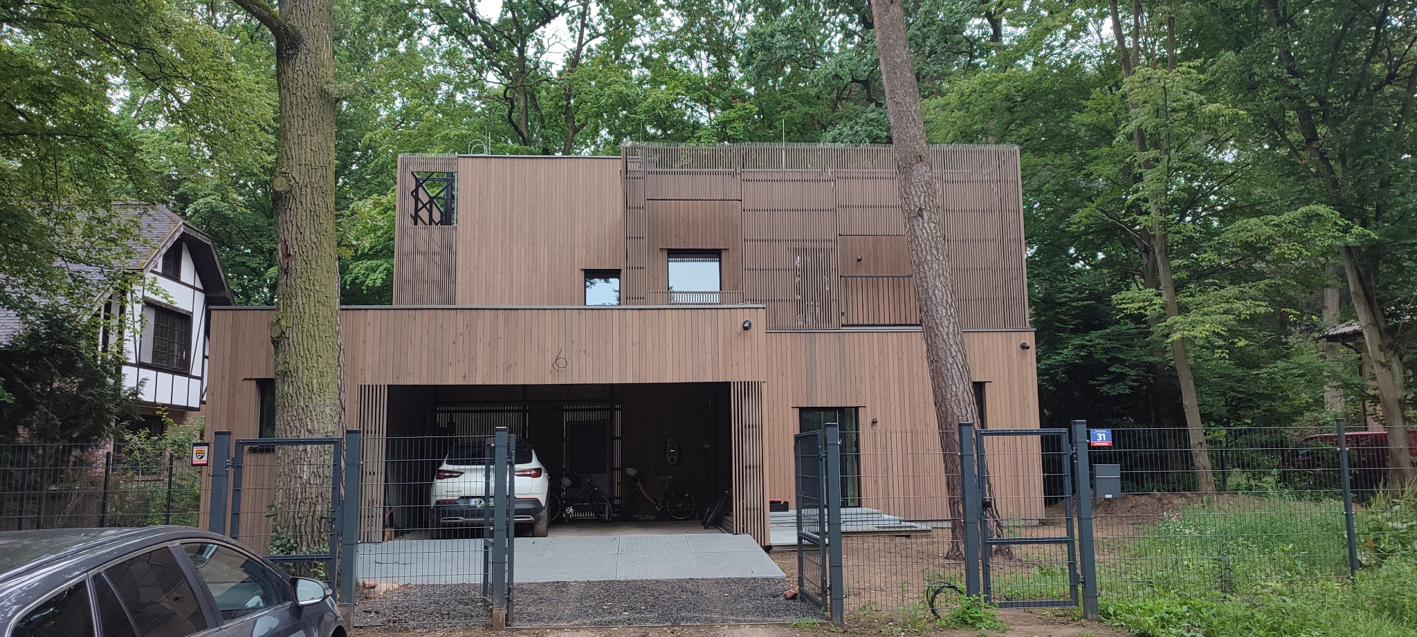 Nowoczesny dom z drewnianą elewacją i garażem, otoczony zielenią. Widoczny fragment ogrodzenia i samochód w garażu. Nietypowe wykończenie elewacji z pionowych listew.