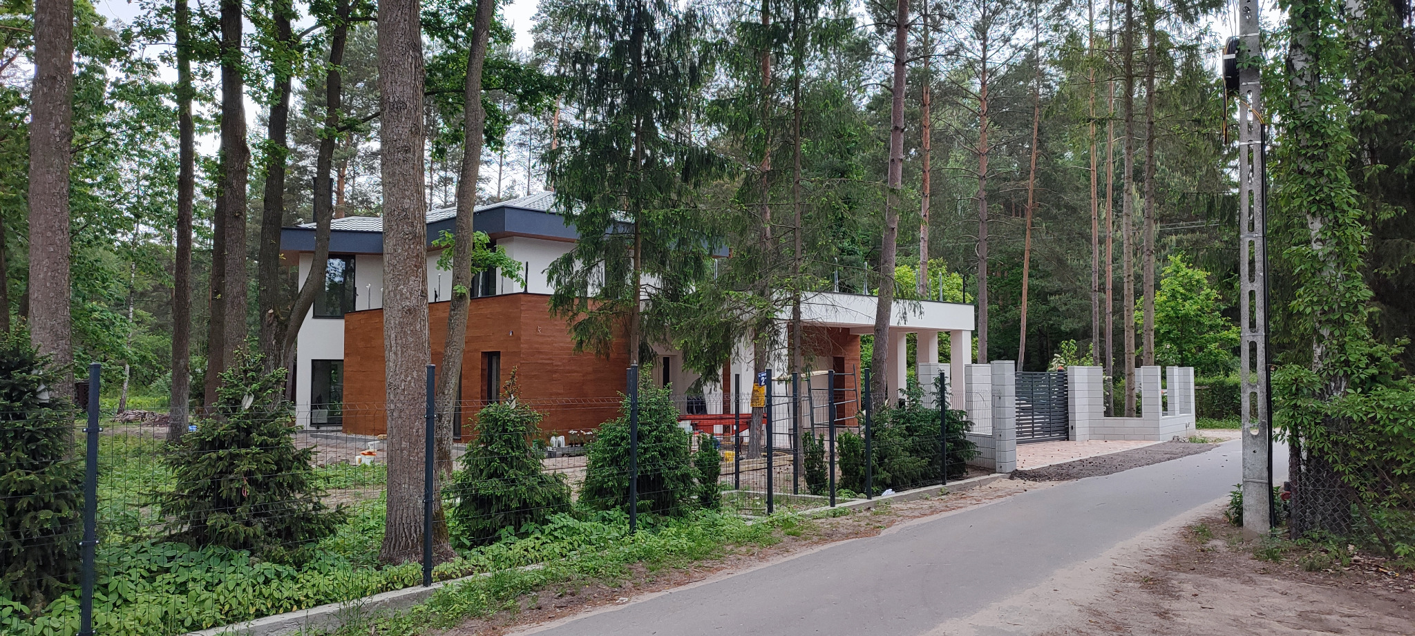 Nowoczesny dom jednorodzinny z drewnianą elewacją, otoczony lasem i ogrodzeniem z bramą wjazdową. Widok z zewnątrz, w tle drzewa i słup energetyczny.