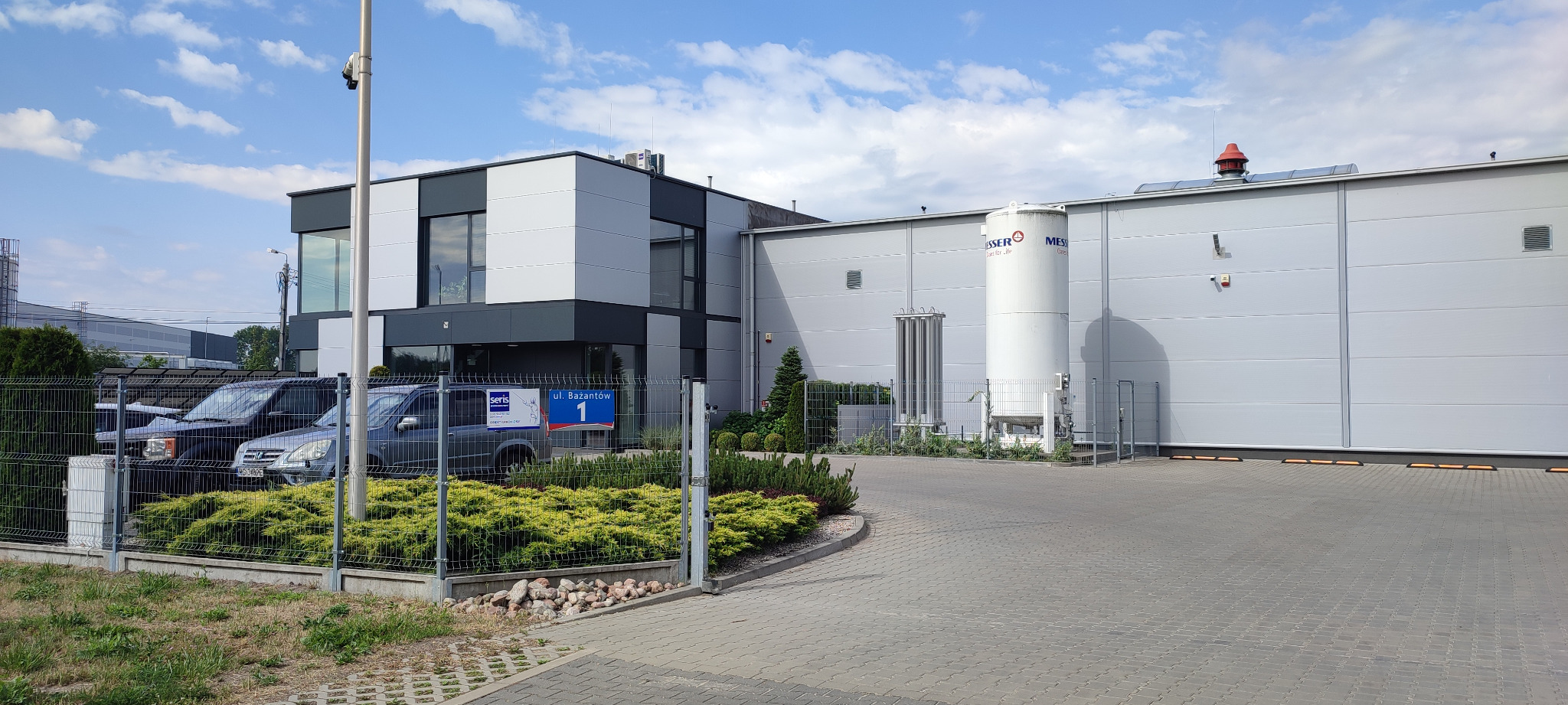 Nowoczesny budynek biurowo-magazynowy z szarą elewacją, widoczne zbiorniki przemysłowe, ogrodzenie z zielenią i zaparkowane samochody. Dzień, błękitne niebo.