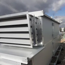 Szary, metalowy system wentylacyjny z żaluzjami na dachu budynku, widoczne elementy montażowe i kamieniste podłoże, pochmurne niebo w tle.