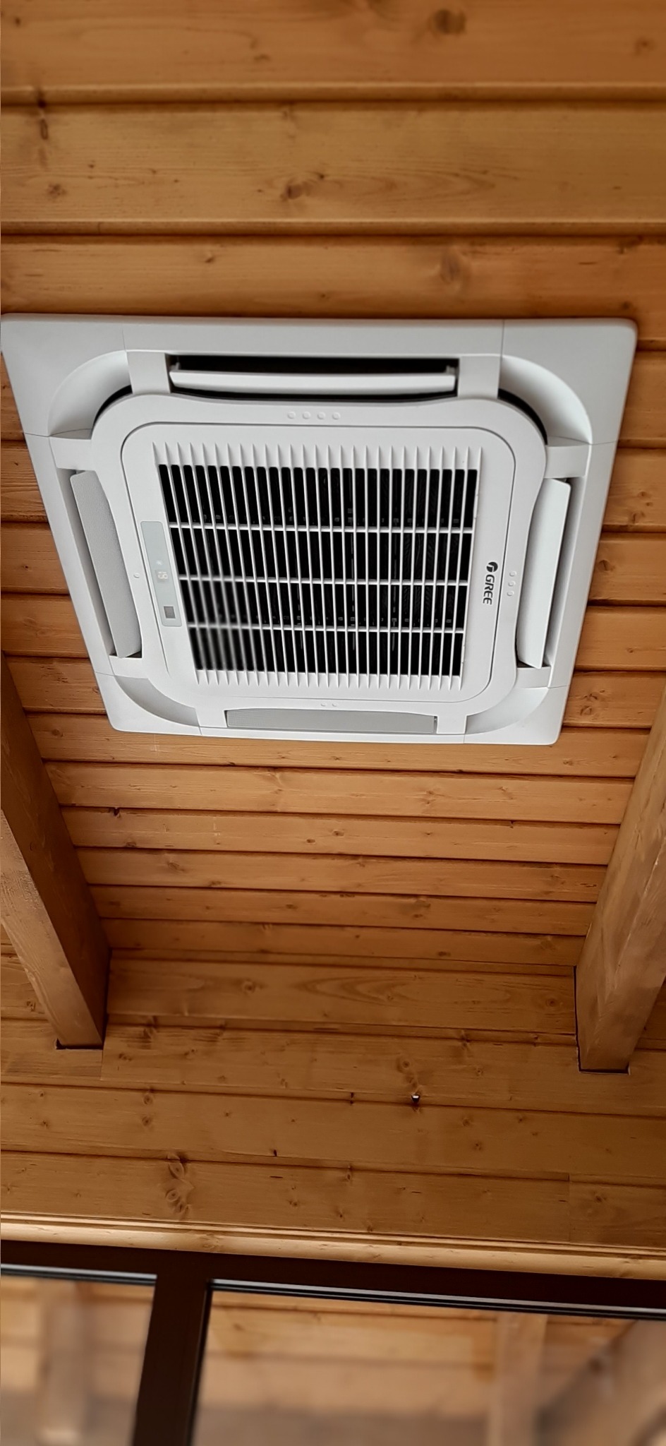 Biała, sufitowa jednostka klimatyzacyjna marki Gree zamontowana w drewnianym pomieszczeniu z widocznymi belkami stropowymi, z perspektywy patrzącego z dołu.
