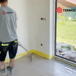 Termolit - Aplikacja wylewki samopoziomującej na ogrzewanie podłogowe, widoczne rury i siatka grzewcza, pracownik w ubrudzonych kaloszach, t-shirt z logo firmy, żółta taśma dylatacyjna przy ścianach.
