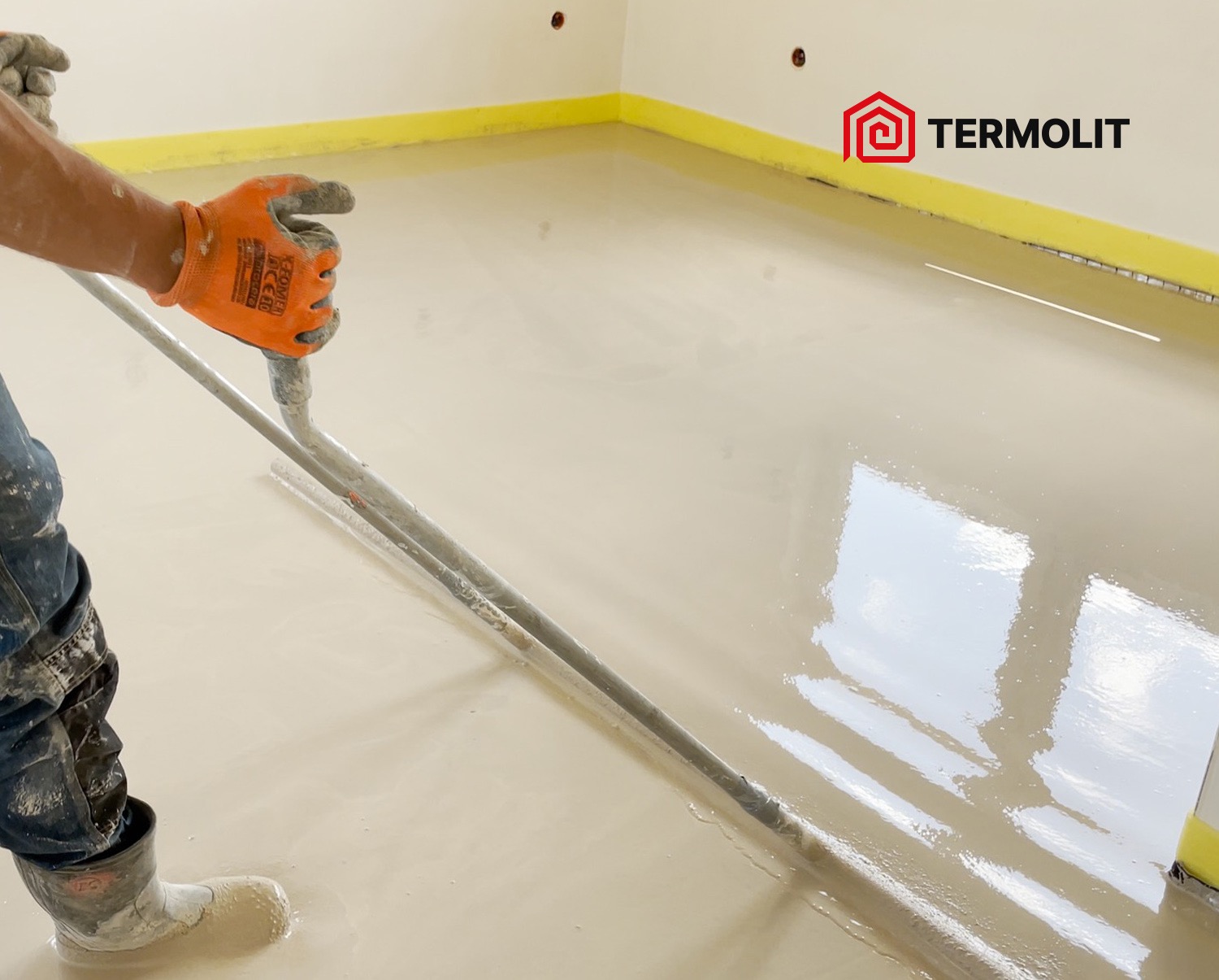Aplikacja samopoziomującej wylewki na podłodze, widoczny pracownik w roboczych ubraniach i pomarańczowych rękawicach rozprowadzający masę za pomocą listwy, logo firmy w prawym górnym rogu.