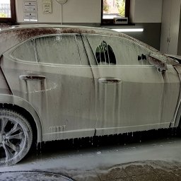 CarsSpa - Biały SUV pokryty pianą myjącą wewnątrz myjni, widoczne półki z kosmetykami samochodowymi w tle.