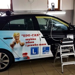 CarsSpa - Czarny samochód oklejony reklamą szkoły nauki jazdy, czyszczony parą za pomocą specjalistycznego urządzenia stojącego na aluminiowej drabinie, w tle widoczne półki z preparatami.