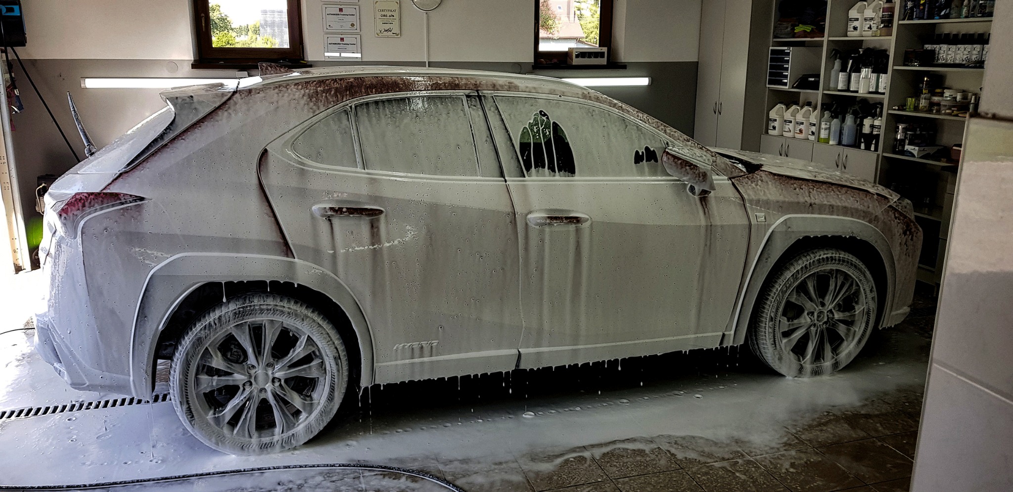 Biały SUV pokryty pianą myjącą wewnątrz myjni, widoczne półki z kosmetykami samochodowymi w tle.