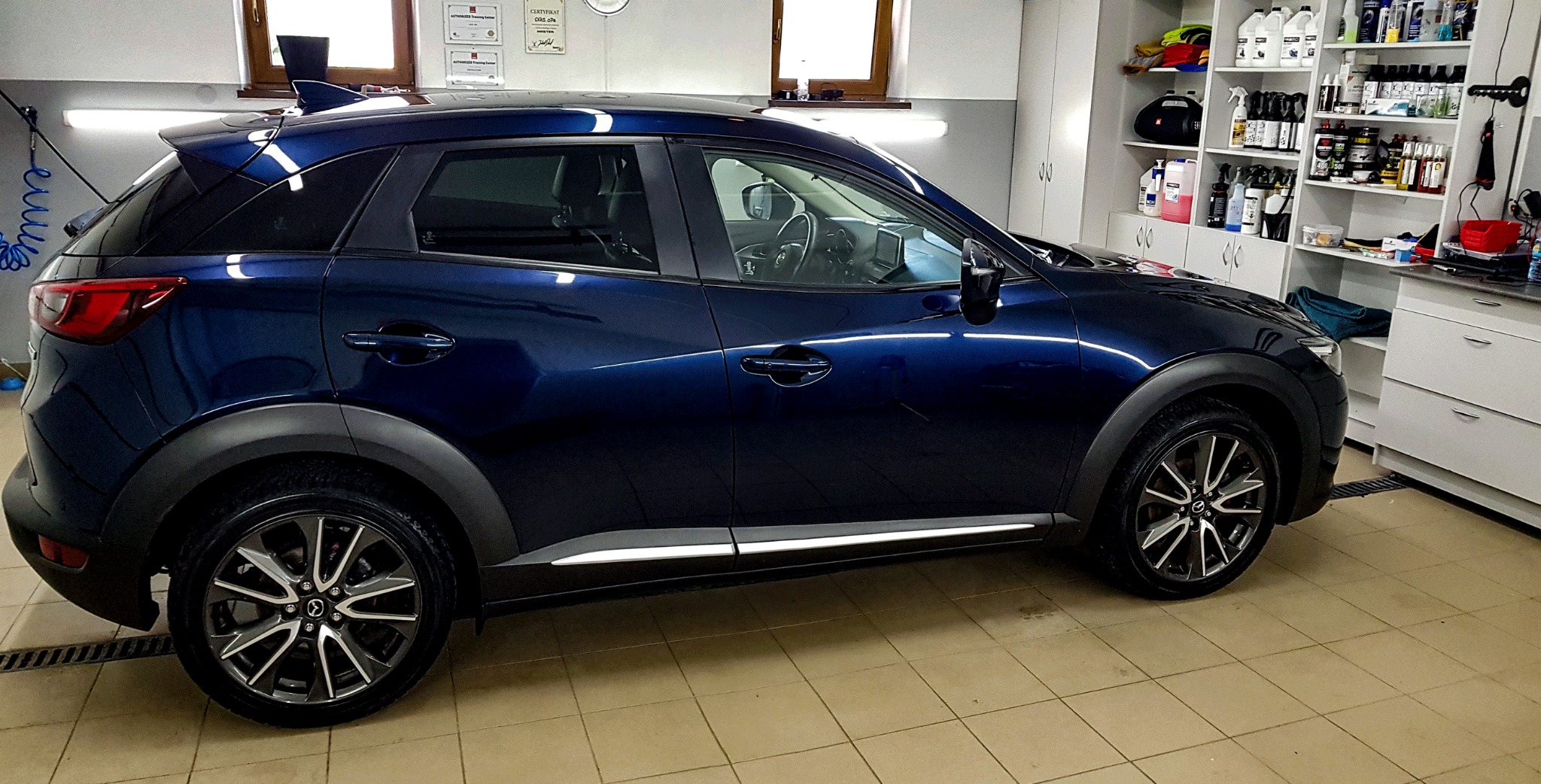 Błyszczący, granatowy samochód typu crossover Mazda CX-3 w jasnym pomieszczeniu z kafelkami na podłodze, widoczne półki z preparatami do pielęgnacji aut i niebieski wąż ciśnieniowy.