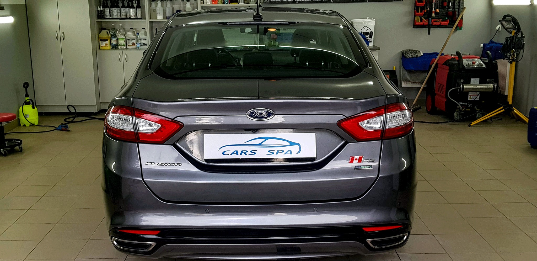 Tył szarego samochodu Ford Fusion w myjni, z widocznym logo Cars Spa na tablicy rejestracyjnej. W tle widoczne półki z detergentami i sprzęt do czyszczenia.