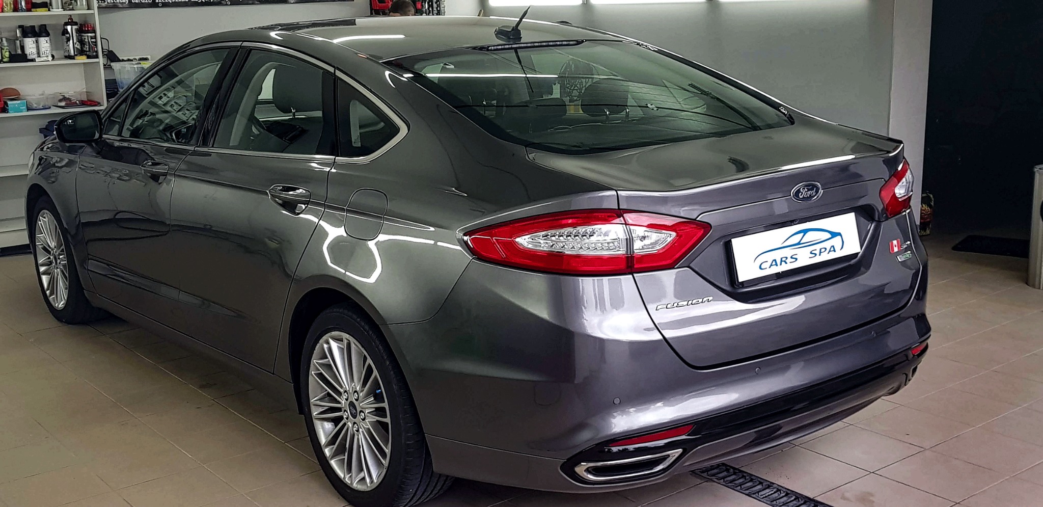 Błyszczący, szary Ford Fusion po usłudze mycia, widoczny z tyłu, z logo Cars Spa na karoserii i odbiciami światła na lakierze.
