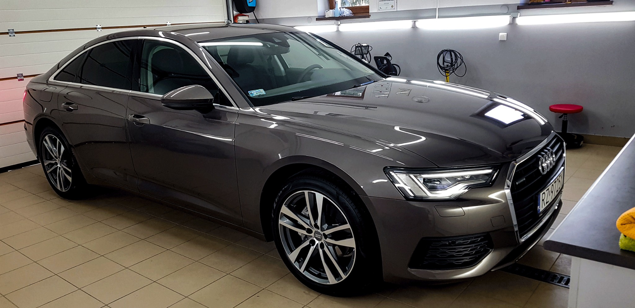 Błyszczący, szary samochód Audi po umyciu w myjni, widoczne odbicia światła na lakierze, wnętrze myjni z białymi drzwiami i szarą ścianą w tle.