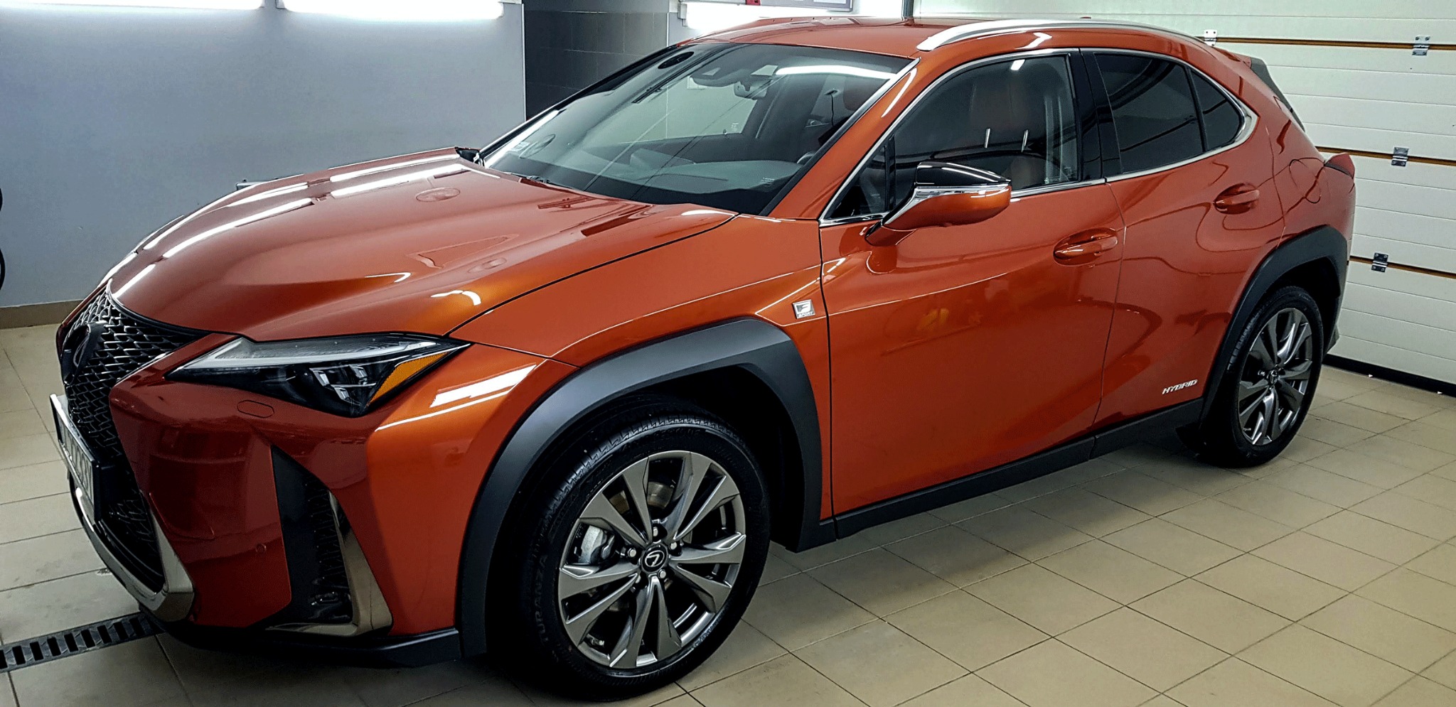 Błyszczący, pomarańczowy Lexus UX Hybrid po umyciu, widoczny z boku na jasnej posadzce myjni, z zamkniętą białą bramą garażową w tle.