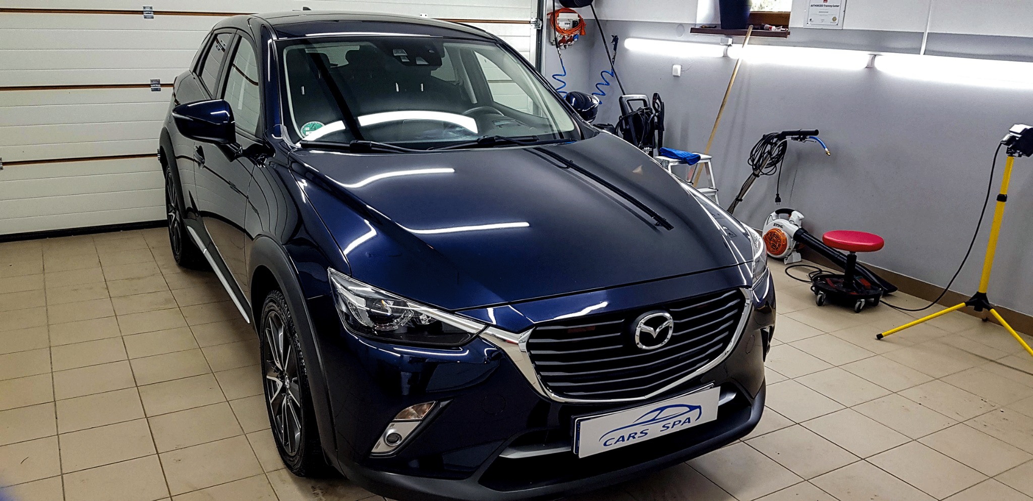 Błyszczący, granatowy samochód Mazda CX-3 po zabiegach pielęgnacyjnych w myjni, z widocznym logo 'Cars Spa' na dolnej części grilla, w tle sprzęt do czyszczenia i oświetlenie warsztatowe.