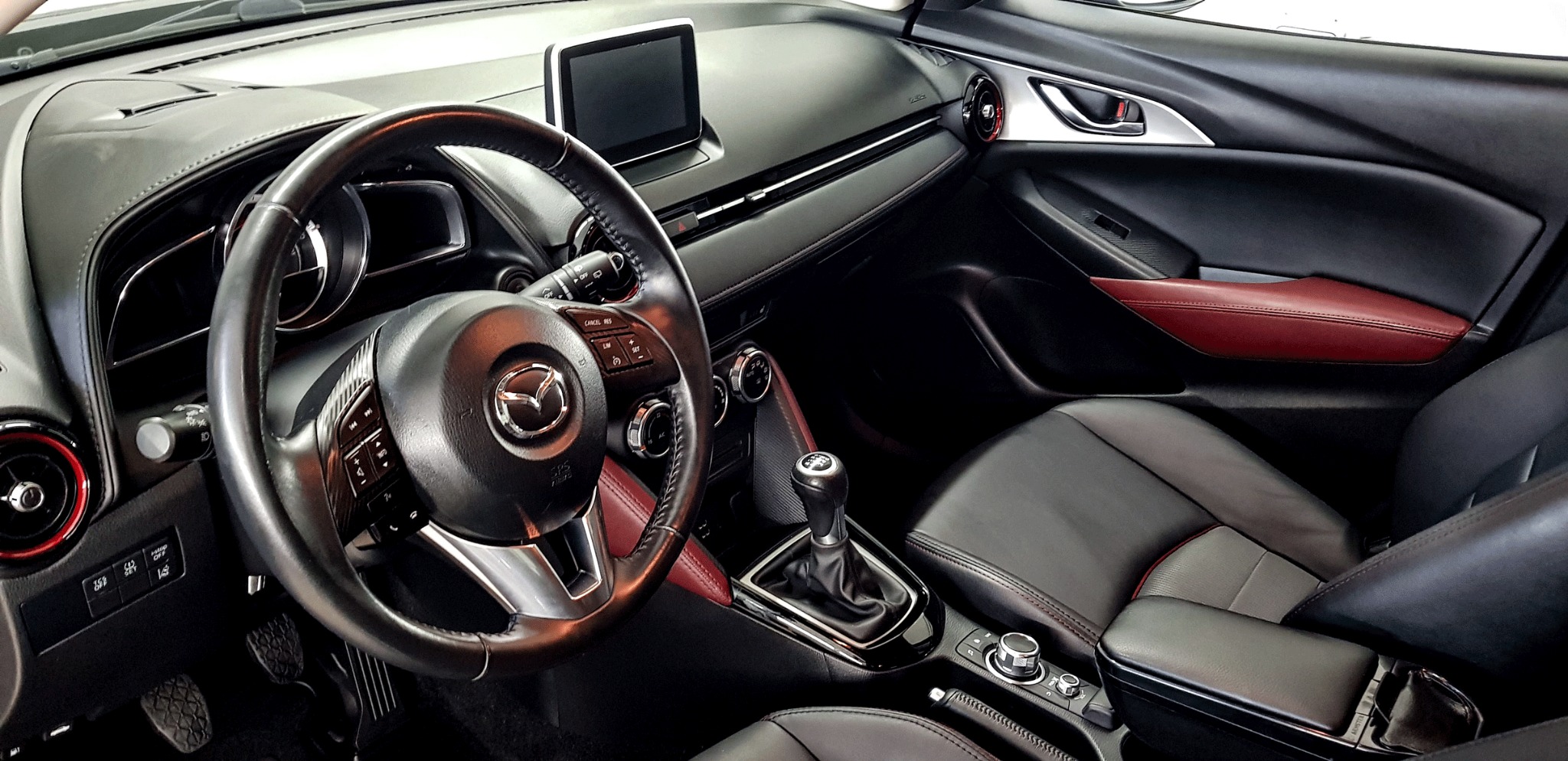 Wnętrze samochodu Mazda z czarną skórzaną tapicerką i czerwonymi akcentami, widok na kierownicę, deskę rozdzielczą i dźwignię zmiany biegów.