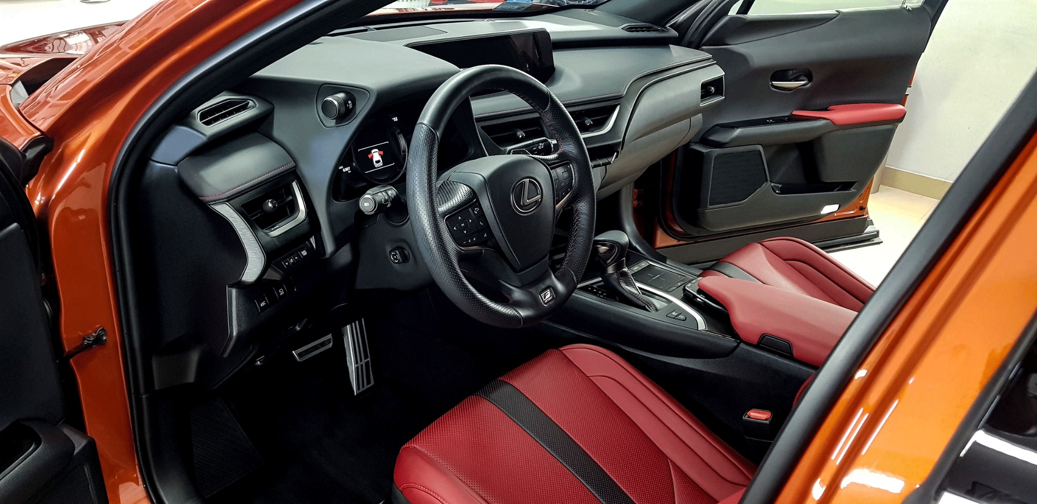 Wnętrze pomarańczowego samochodu Lexus z czarną deską rozdzielczą i czerwonymi skórzanymi siedzeniami, widok z otwartych drzwi kierowcy.