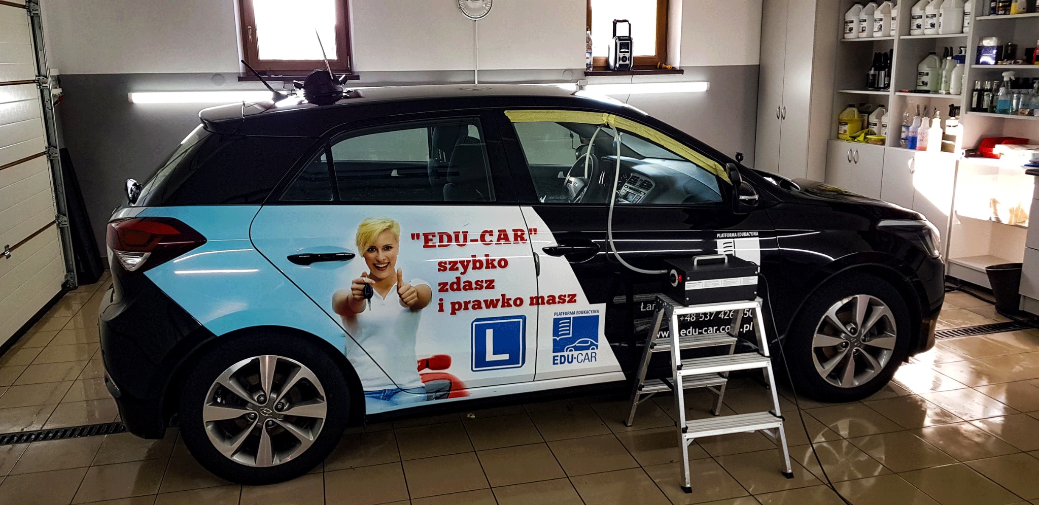 Czarny samochód oklejony reklamą szkoły nauki jazdy, czyszczony parą za pomocą specjalistycznego urządzenia stojącego na aluminiowej drabinie, w tle widoczne półki z preparatami.
