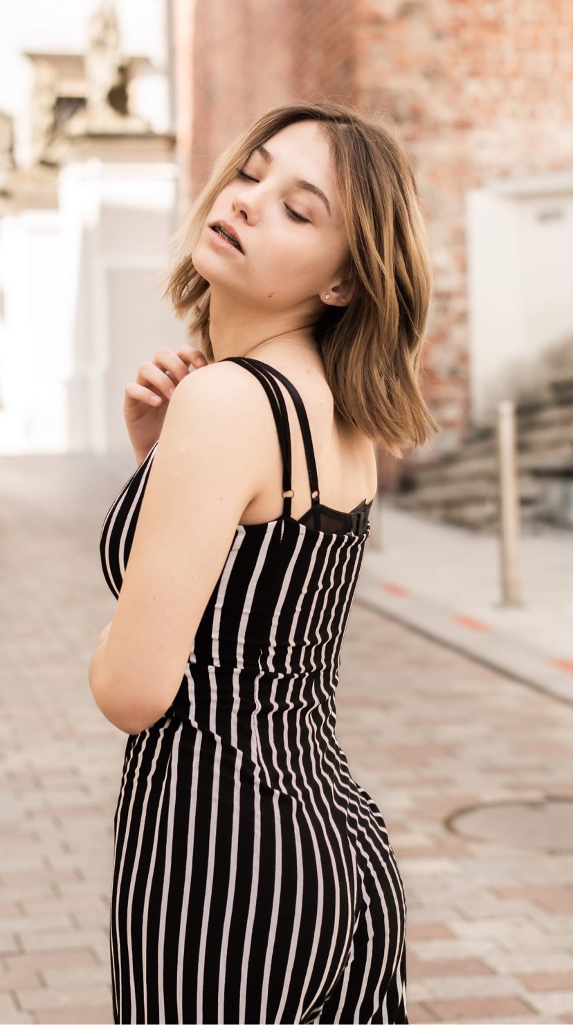 Portret młodej kobiety w kombinezonie w paski, pozującej na tle ceglanego budynku i brukowanej uliczki.