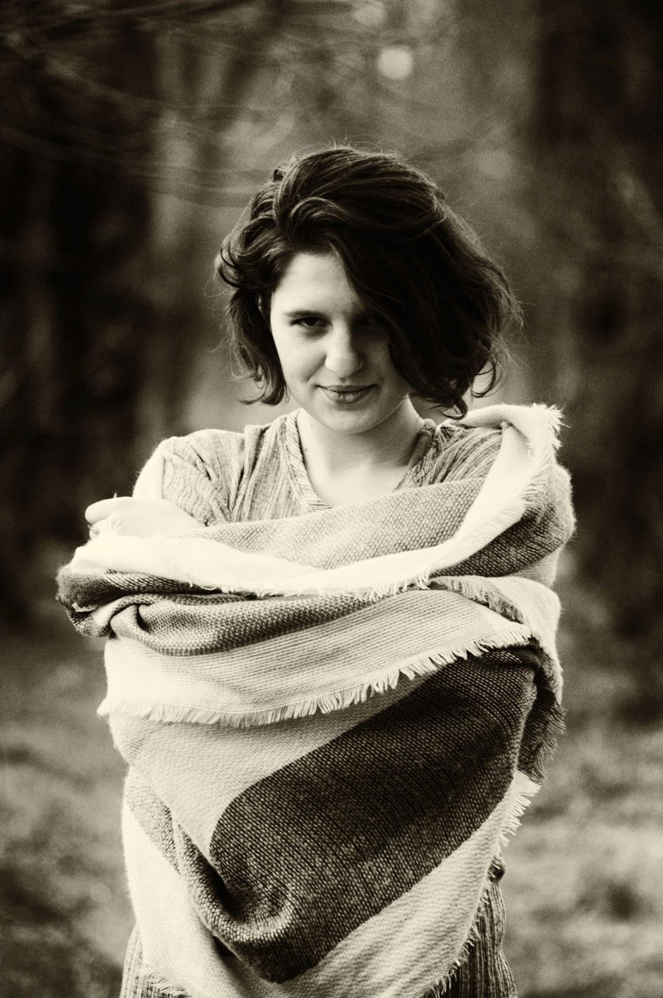 Portret kobiety w Szczecinie, owiniętej w gruby, pasiasty koc z frędzlami, na rozmytym tle drzew w sepii.