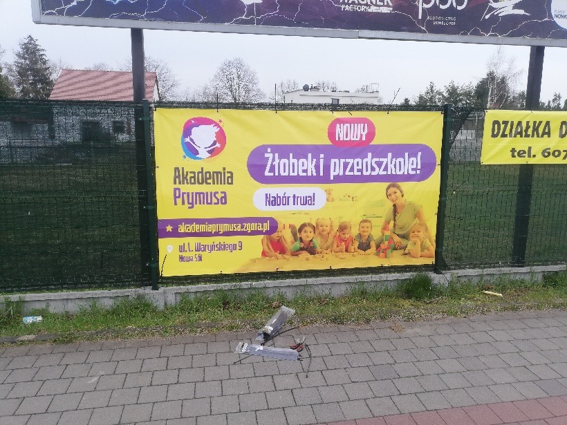 Żółty baner reklamowy żłobka i przedszkola Akademia Prymusa z uśmiechniętymi dziećmi i informacją o naborze, zawieszony na zielonym ogrodzeniu, z butelką i śmieciami na chodniku przed nim.