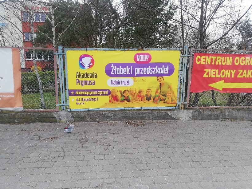 Żółty baner reklamowy na ogrodzeniu siatkowym, promujący Akademię Prymusa: żłobek i przedszkole, z adresem strony internetowej i adresem stacjonarnym w Nowej Soli.