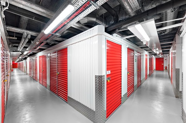 Korytarz z rzędami czerwonych i białych boksów do wynajęcia, zabezpieczonych kłódkami, widok z perspektywy, rury wentylacyjne pod sufitem.