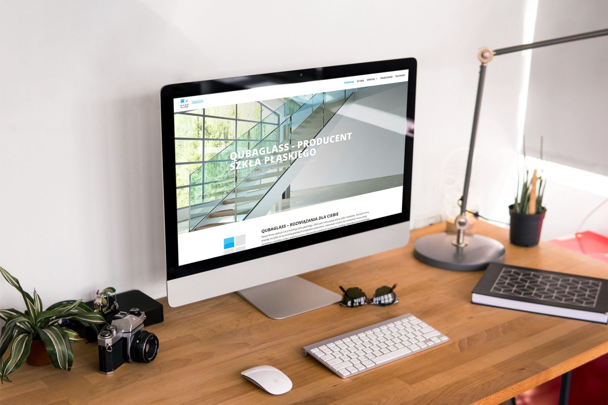 Monitor wyświetla stronę internetową firmy Qubaglass, producenta szkła płaskiego, na biurku z rośliną, aparatem analogowym, okularami i lampką.
