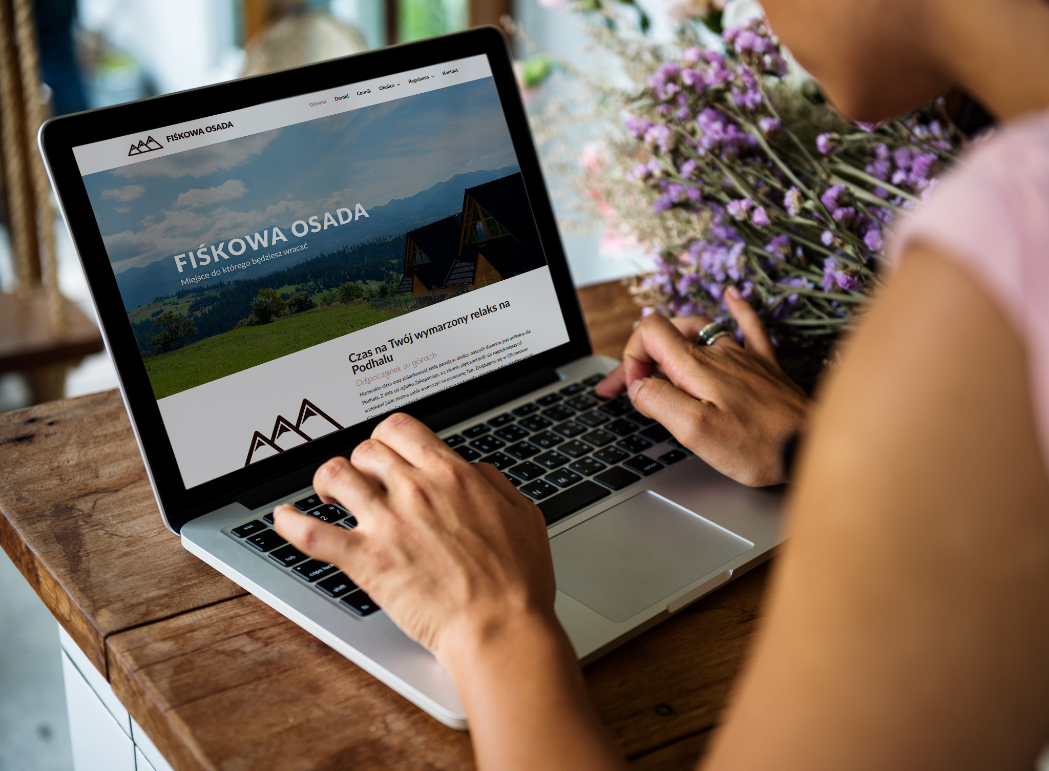 Kobieta przegląda stronę internetową Fiskowaosada.pl na laptopie, obok bukiet fioletowych kwiatów, widoczny fragment drewnianego blatu.