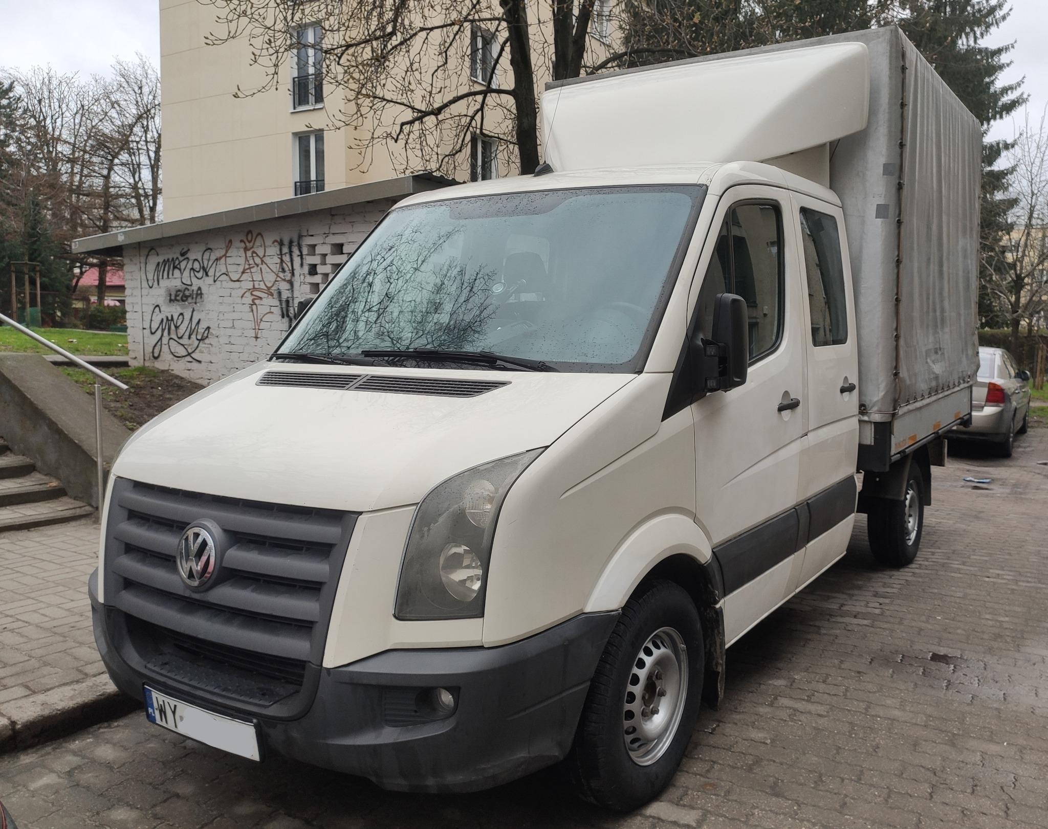 Biały samochód dostawczy Volkswagen Crafter z plandeką na parkingu miejskim, widoczne graffiti na ścianie w tle.