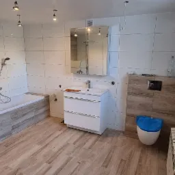 Ukończona łazienka z białymi płytkami ściennymi, drewnopodobną podłogą, kabiną prysznicową typu walk-in, białą szafką z umywalką i toaletą z niebieską deską, widoczne wiszące kable oświetleniowe.