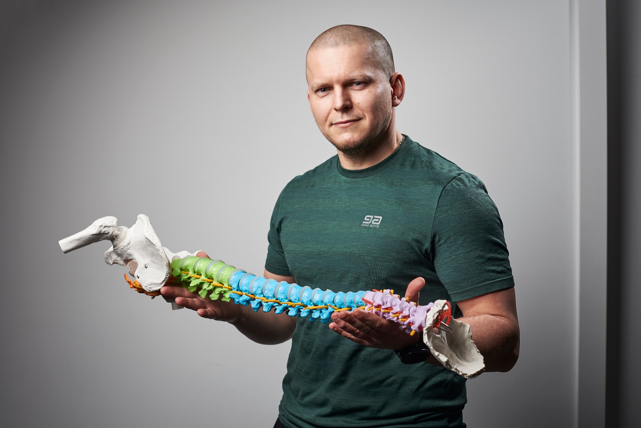 Mężczyzna w zielonej koszulce trzyma model kręgosłupa z miednicą, prezentacja edukacyjna lub demonstracja terapeutyczna.