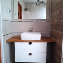 Nowoczesna łazienka w Miłakowie z drewnopodobnymi płytkami, białą umywalką nablatową na drewnianym blacie i szafką pod umywalką z dwoma szufladami oraz lustrem, w którym odbija się sufit...