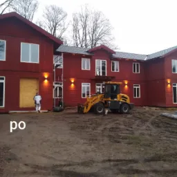 Czerwony, dwukondygnacyjny budynek z szarą dachówką w trakcie prac wykończeniowych, z pracownikiem w białym kombinezonie przykrywającym okno płytą OSB, żółtą ładowarką WOLF i stertą metalowych...