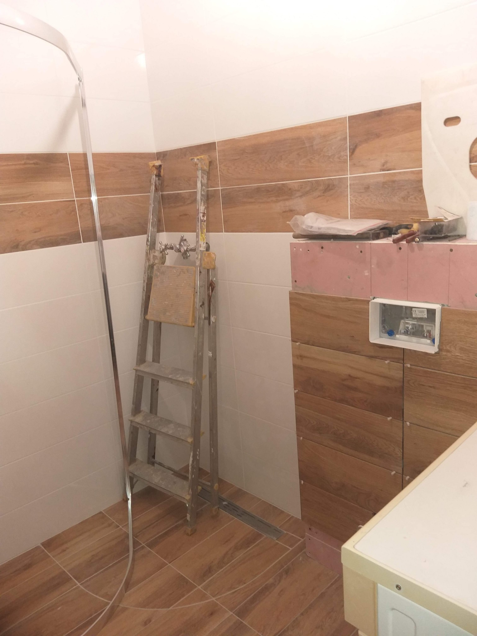Łazienka w trakcie wykańczania: widoczne płytki imitujące drewno na ścianach i podłodze, drabina z narzędziami, zabudowa podtynkowego systemu sanitarnego z różową płytą karton-gips.