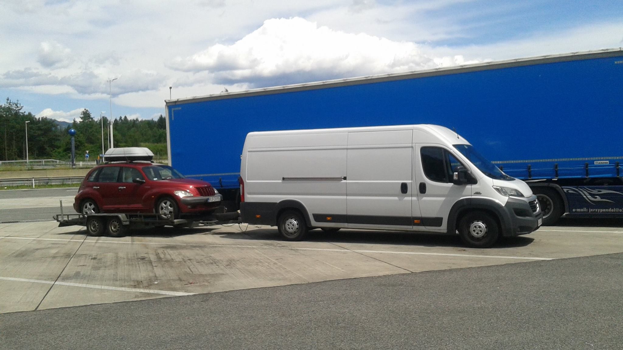 Czerwony samochód osobowy z bagażnikiem na dachu, transportowany na przyczepie holowanej przez biały samochód dostawczy na tle niebieskiej naczepy ciężarówki i pochmurnego nieba.