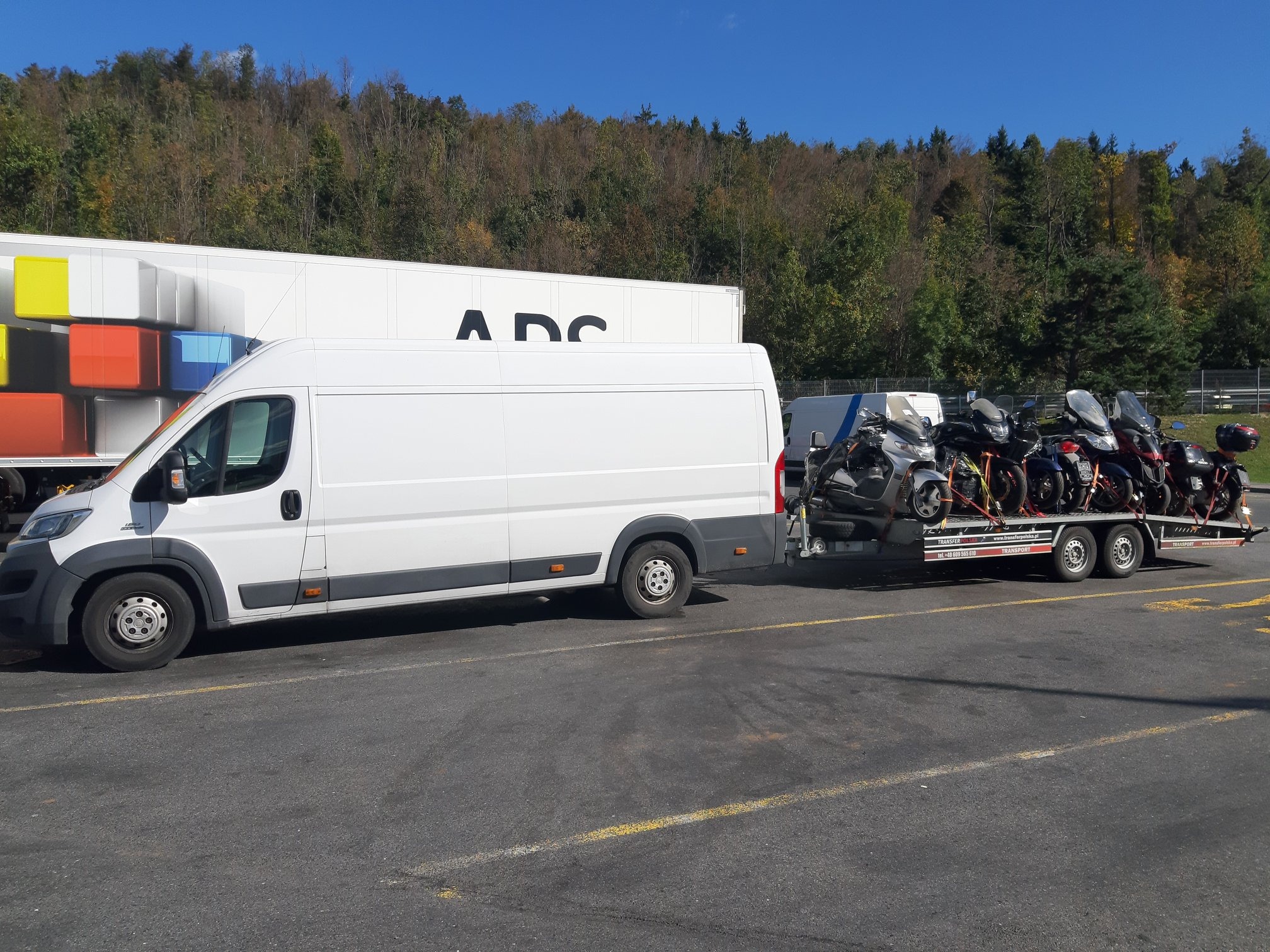 Biały furgon z przyczepą przewożącą motocykle na tle lasu i naczepy z kolorowymi elementami.