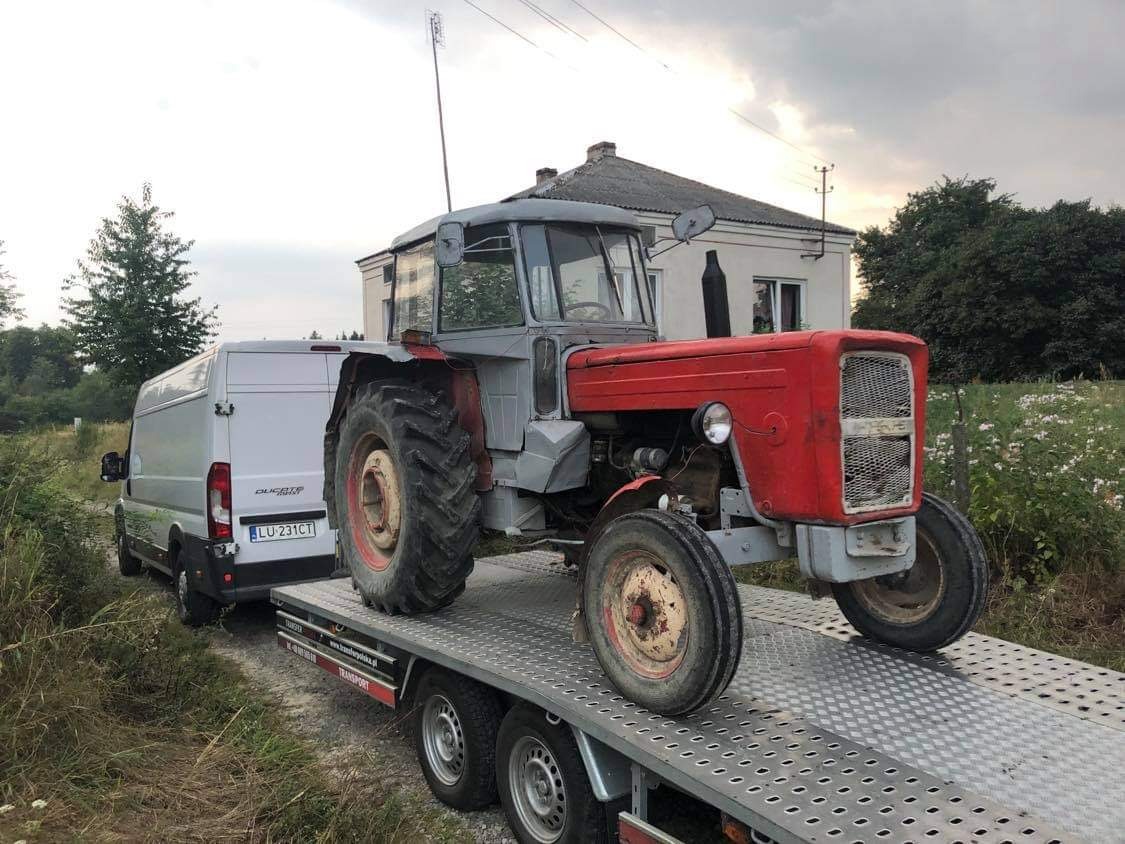 Czerwono-szary traktor Ursus na lawecie transportowej, ciągniętej przez biały bus Ducato z polskimi tablicami rejestracyjnymi, na tle wiejskiego krajobrazu z domem.