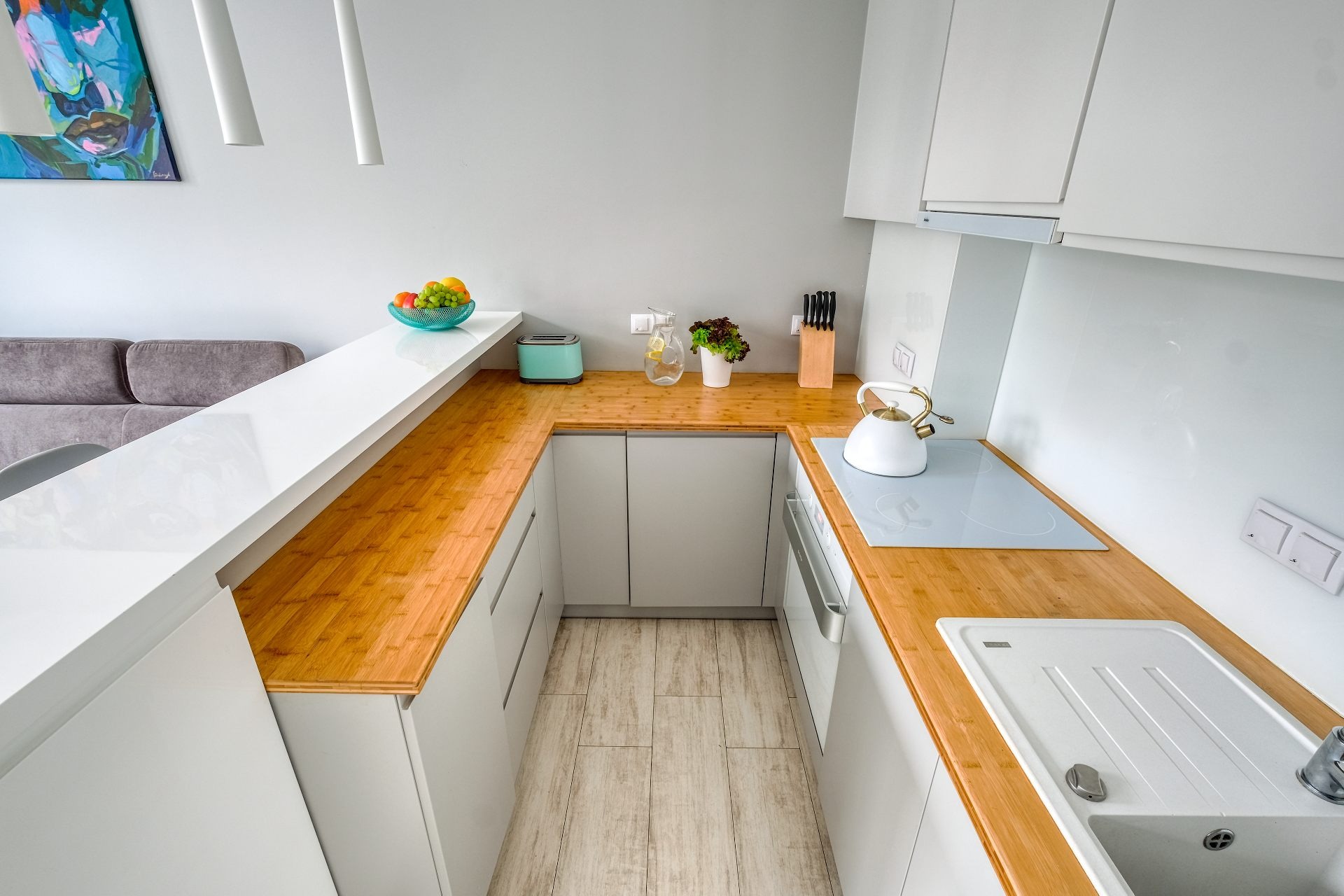 Nowoczesna kuchnia z drewnianym blatem, białymi szafkami i płytą indukcyjną, uzupełniona o czajnik w stylu retro i misę z owocami