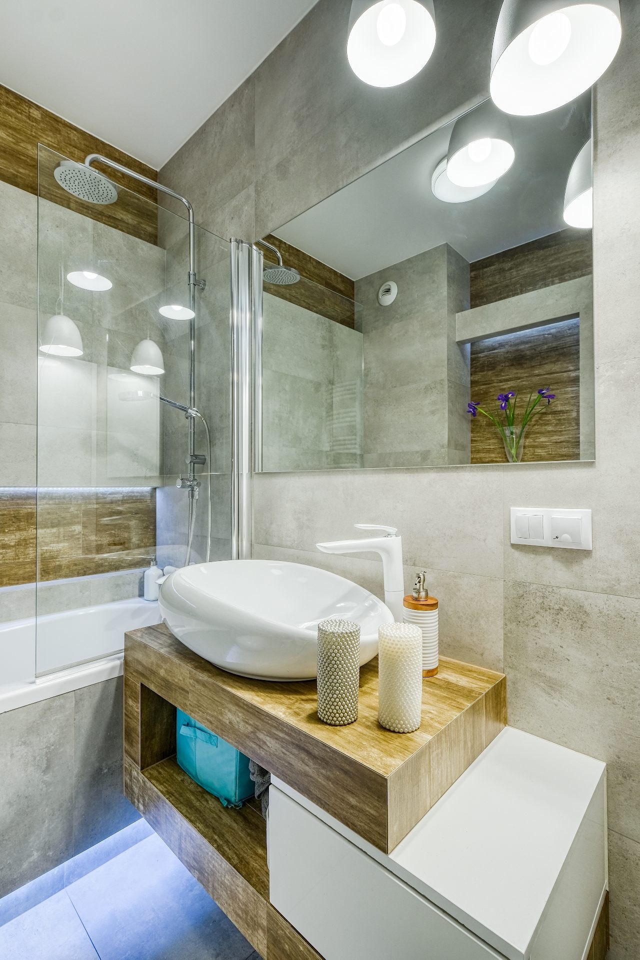 Nowoczesna łazienka z owalną umywalką nablatową, drewnopodobnym blatem i szarą płytką, oświetlona punktowymi lampami sufitowymi, z widoczną kabiną prysznicową i dekoracyjnymi świecami.