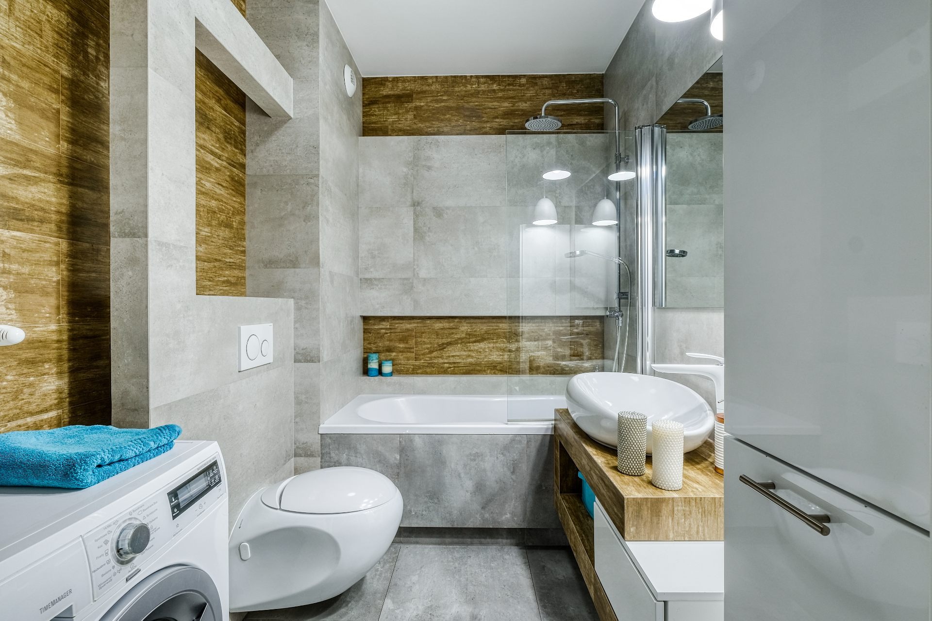 Nowoczesna łazienka z szarymi i drewnopodobnymi płytkami, wolnostojącą umywalką na drewnianym blacie, toaletą typu Geberit i pralką z turkusowym ręcznikiem.