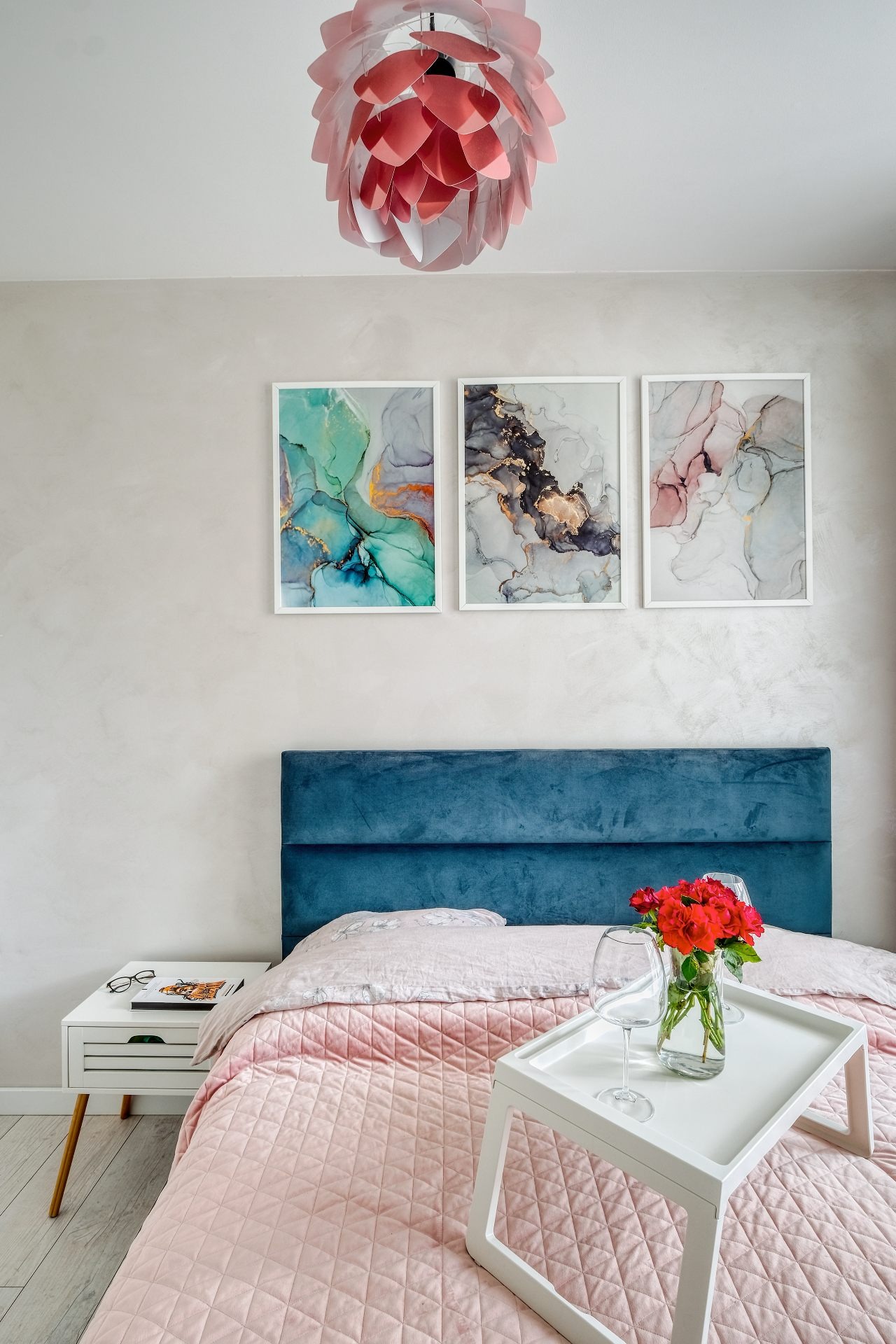 Sypialnia w pastelowych kolorach z niebieskim zagłówkiem, trzema abstrakcyjnymi obrazami na ścianie, lampą w kształcie kwiatu i śniadaniem na łóżku.