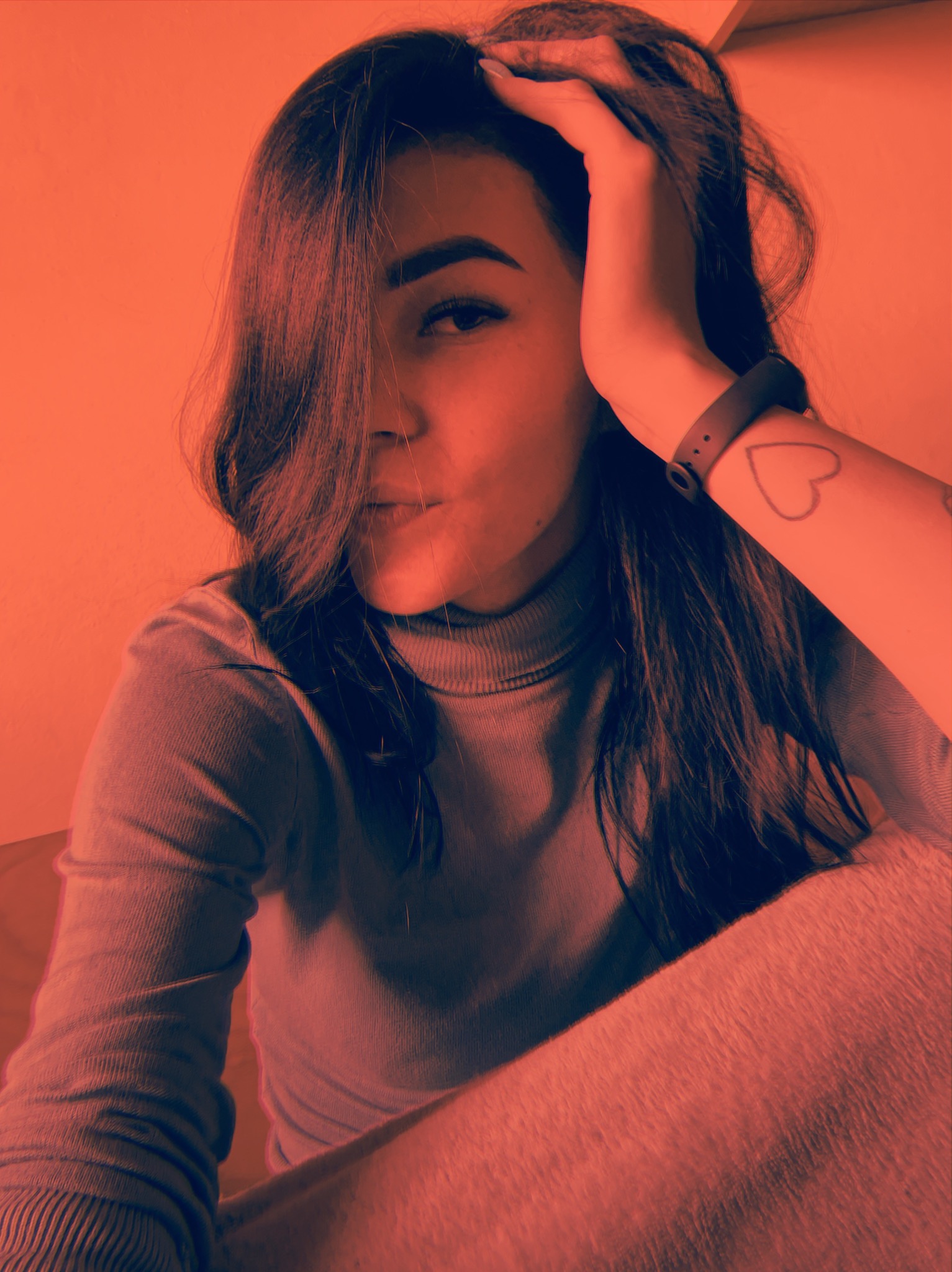 Portret kobiety z ręką we włosach, widoczny tatuaż serca na przedramieniu i smartwatch na nadgarstku, włosy opadają na twarz, stonowana kolorystyka.