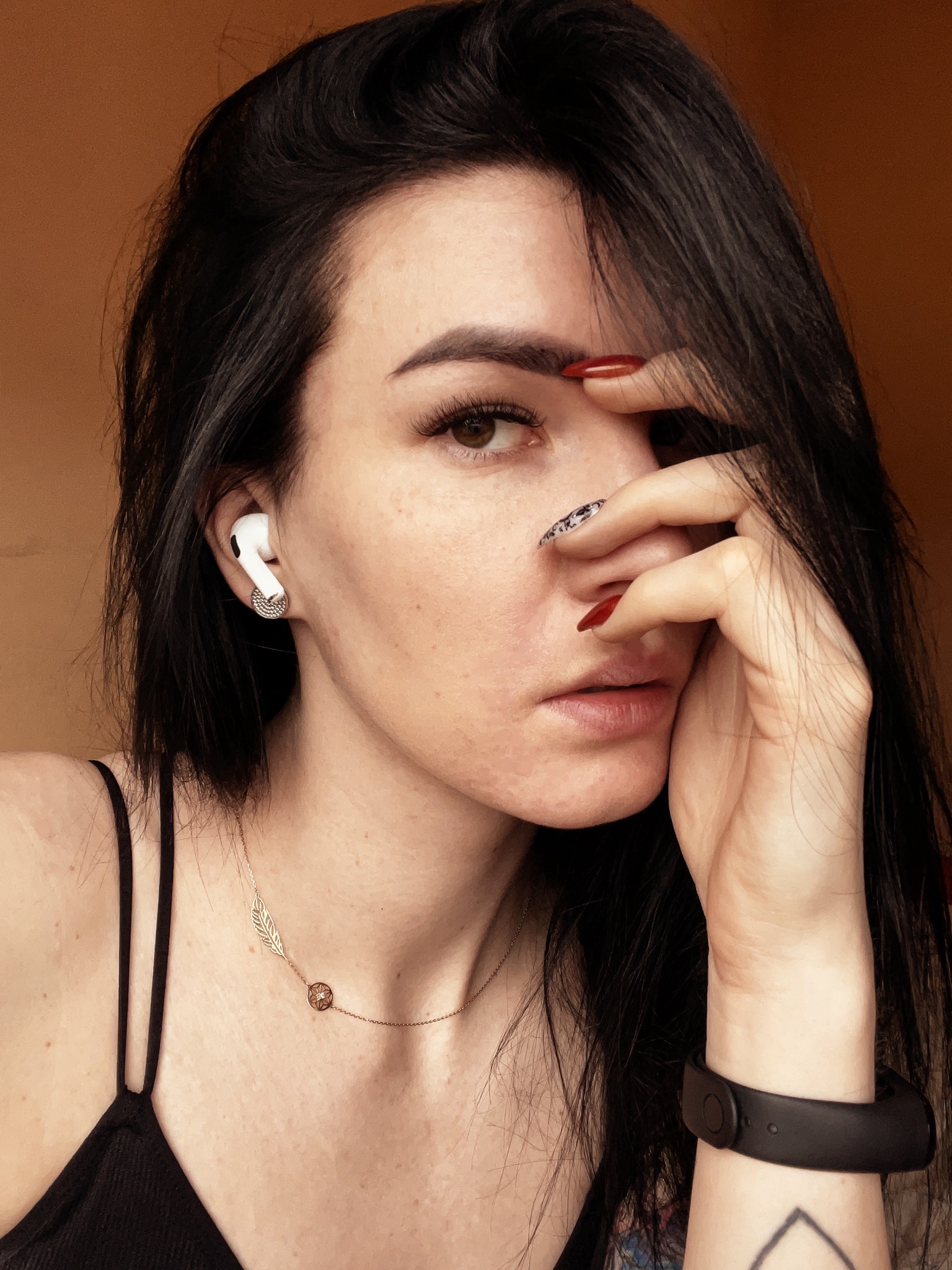 Portret kobiety z ciemnymi włosami, zasłaniającej twarz dłonią z czerwonym manicure, noszącej słuchawkę bezprzewodową i złotą biżuterię.