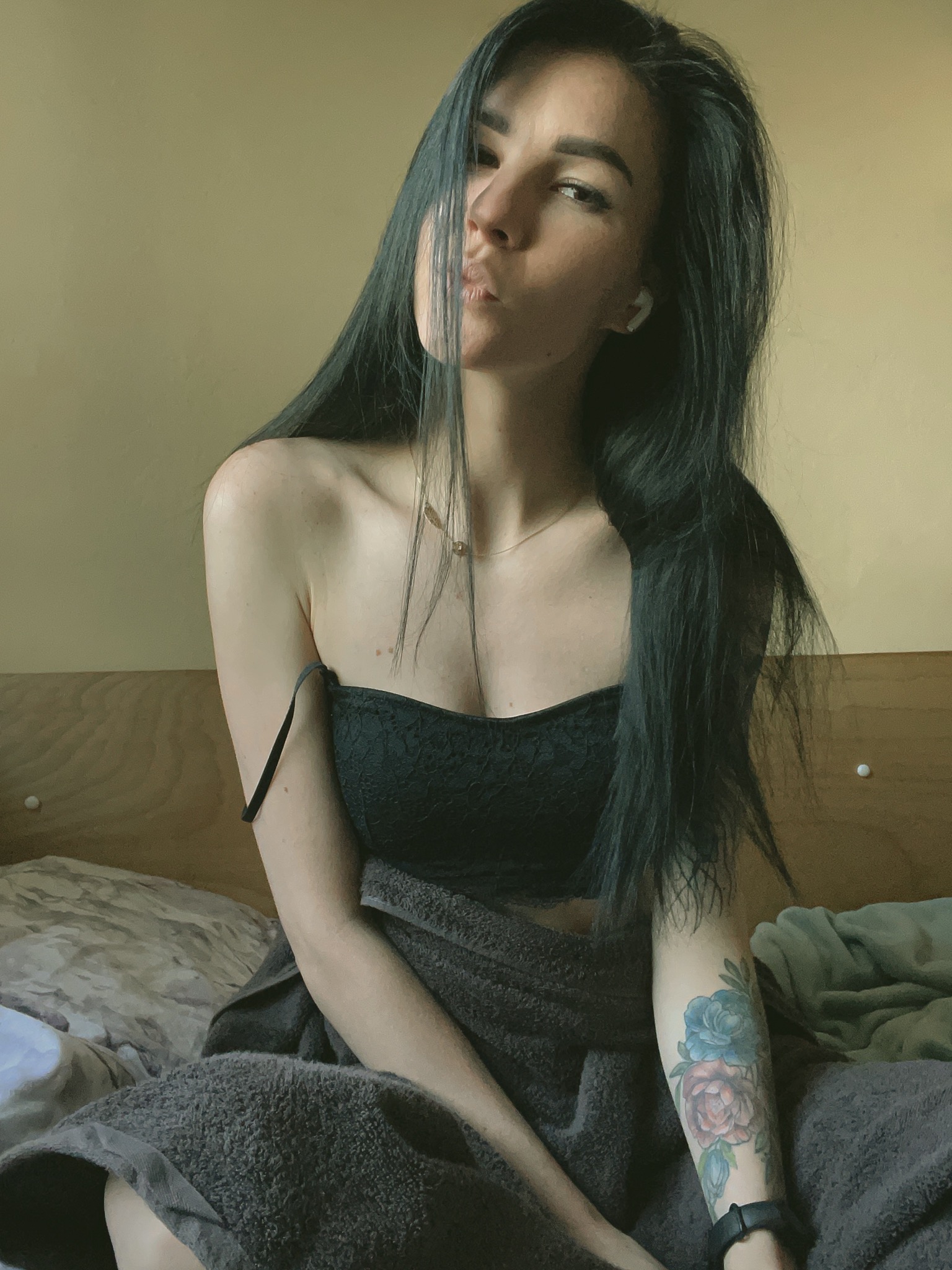 Kobieta z tatuażem kwiatowym na ramieniu, czarnym topie i słuchawką bezprzewodową w uchu, siedząca na łóżku z szarą narzutą, robiąca dzióbek.