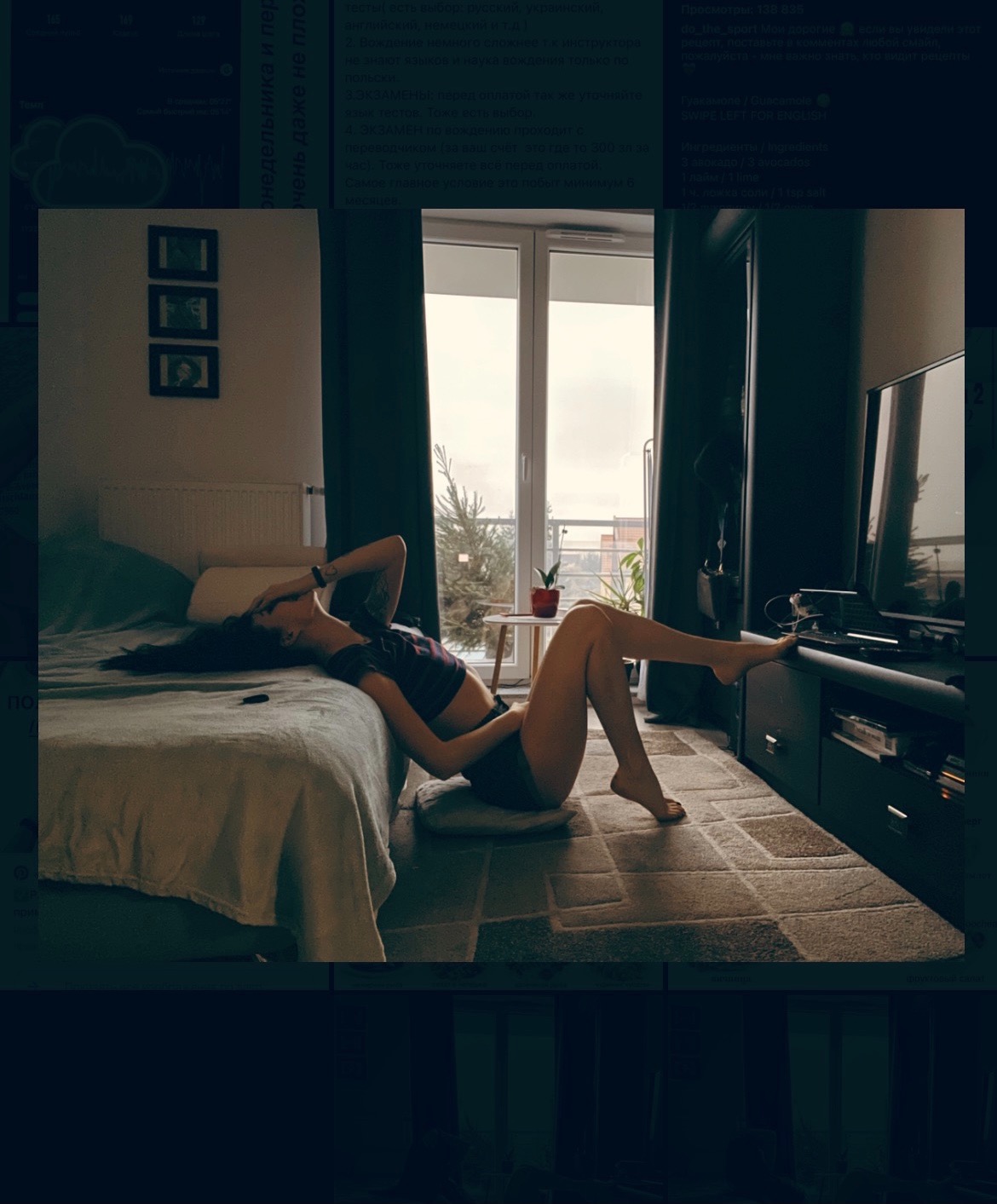 Kobieta leżąca na łóżku, opierająca się o poduszkę na podłodze, z widokiem na okno i balkon, w tle telewizor i meble w ciemnych kolorach.