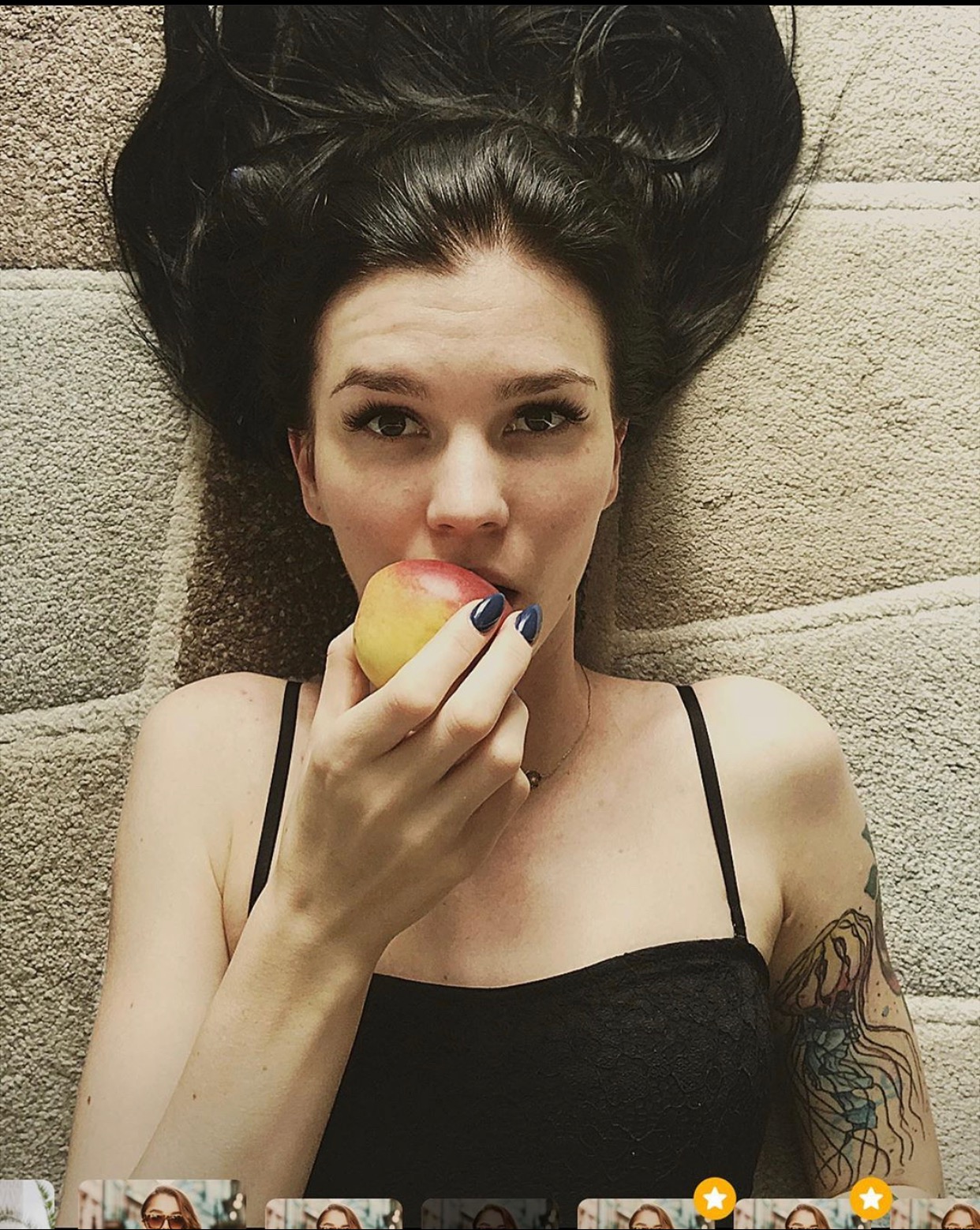 Kobieta z tatuażem meduzy na ramieniu, leżąca na dywanie, trzyma jabłko w dłoniach z ciemnymi paznokciami, patrząc w obiektyw.
