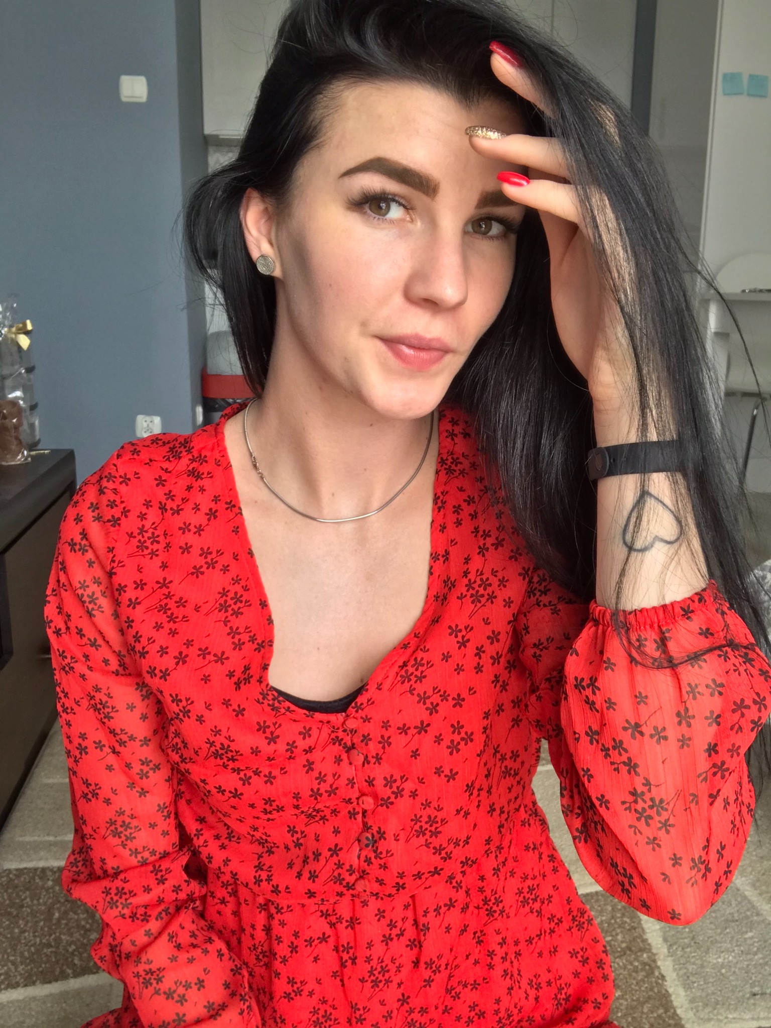 Portret kobiety z długimi, czarnymi włosami, w czerwonej sukience w drobne kwiatki, z tatuażem serca na nadgarstku, spoglądającej w bok, z dłonią uniesioną do czoła.