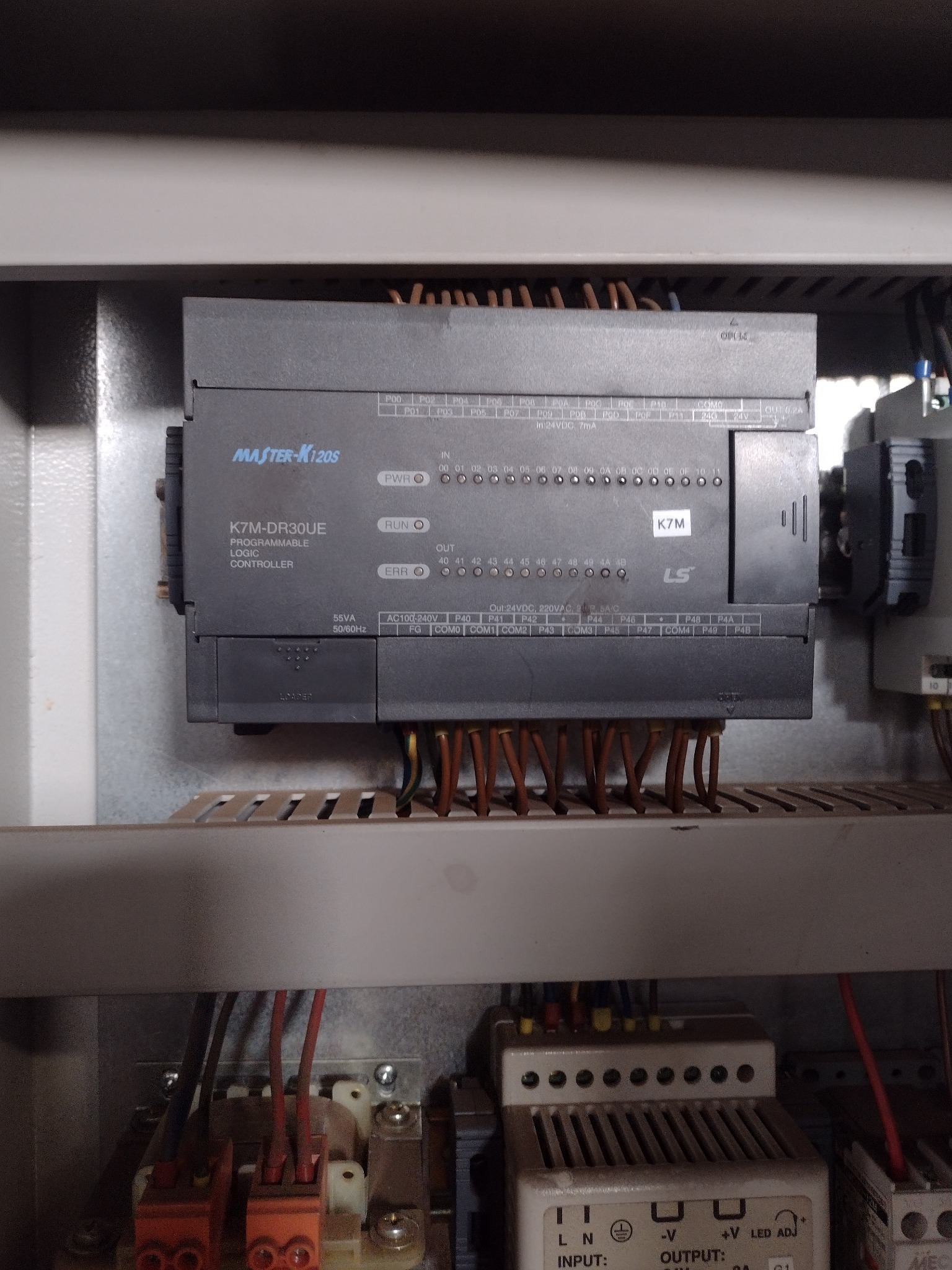 Szary sterownik logiczny programowalny MASTER-K120S, model K7M-DR30UE, zamontowany w szafie sterowniczej z widocznymi przewodami i innymi elementami automatyki.