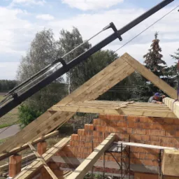 Montaż drewnianej konstrukcji dachu z użyciem dźwigu na murze z czerwonej cegły, w tle drzewa i błękitne niebo.