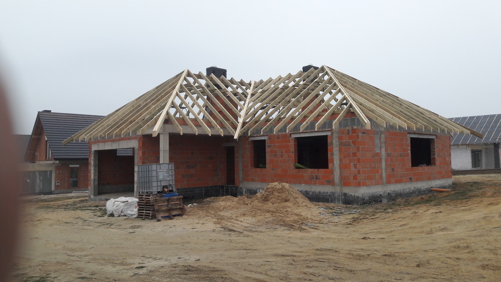 Surowy stan budynku z czerwonej cegły z widoczną drewnianą konstrukcją szkieletową dachu w trakcie budowy, widoczne palety i zbiornik na wodę. W tle dom z pokrytym dachem.