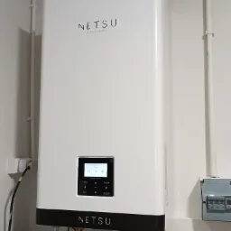 Biała, nowoczesna pompa ciepła marki Netsu, zamontowana na białej ścianie, z widocznym panelem sterowania i podłączeniami hydraulicznymi, w tle skrzynka elektryczna.